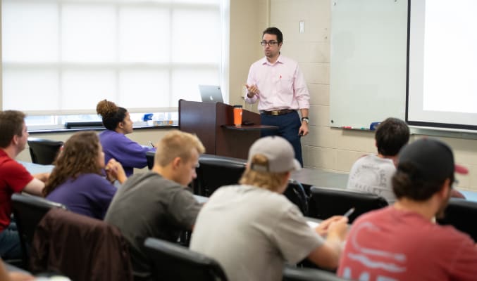 Male professor with glasses presents to a class of students sitting in rows in an academic building on campus.