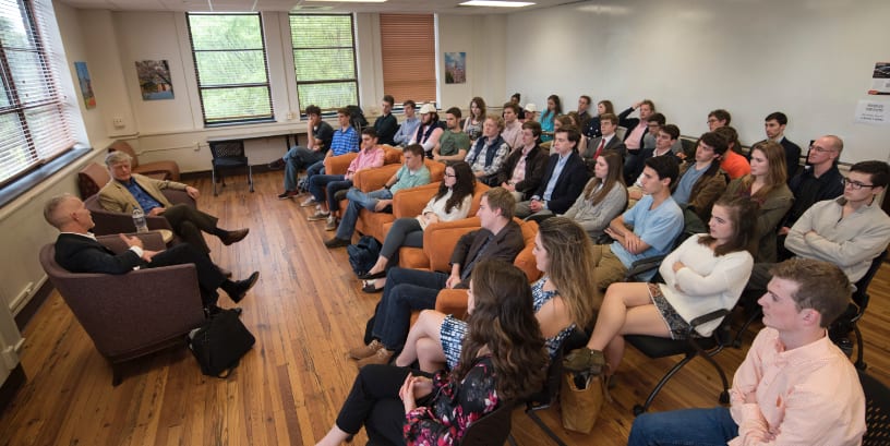 Four rows of seated Clemson students in business attire listen to two men at the front of the classroom.