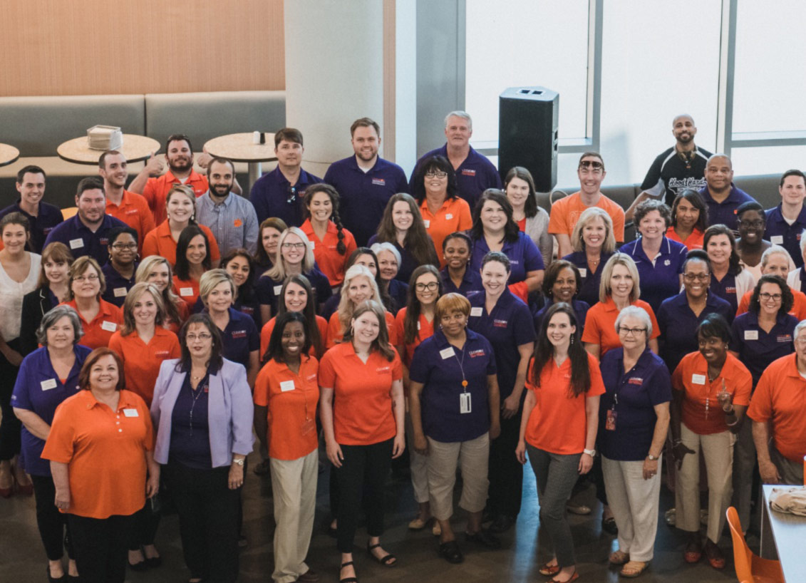 A group of Clemson employees wearing orange and purple pose together, smiling.