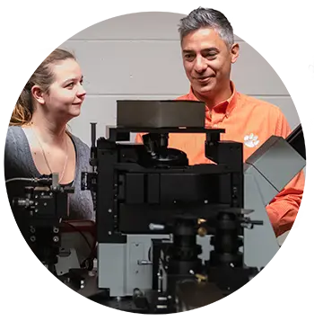 A male professor wearing a striped orange shirt with a white Tiger Paw and a female student look on at a piece of laboratory machinery as a male student looks through a binocular microscope.