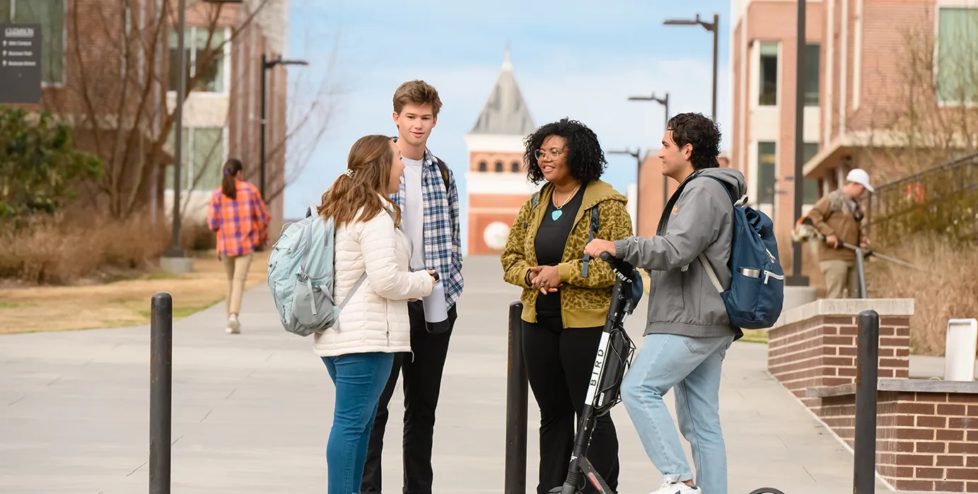 Two male students and two female students stand on a walking path and talk with each other. The top of a brick clock tower building is visible in the distant background. 