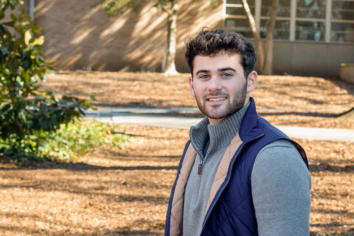 A male student wearing a gray quarter-zip sweater and a blue outerwear vest with a tan interior lining poses for a photo outdoors on campus.