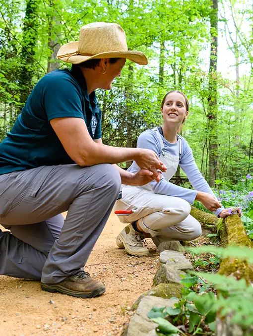 A female student wearing a purple long-sleeved shirt and khaki overalls and a female instructor wearing gray pants, blue-green polo and beige straw hat kneel down next to a garden path with purple flowers and a moss-covered branch. 