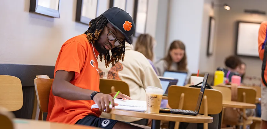 A male student in an orange t-shirt and Clemson hat studies with a notebook and laptop in an on-campus Starbucks.