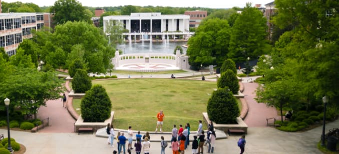 An aerial view of a tour guide addressing his group with the ampitheater, reflection pond and Cooper Library behind him.