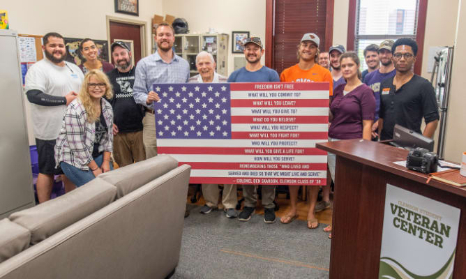A group of staff and students stand behind a flag and smile in the student veteran resource center.