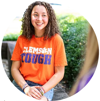 A female student wearing denim shorts and an orange T-shirt that says “Clemson Tough” in white and purple letters sits on a brick ledge and talks to a female student.