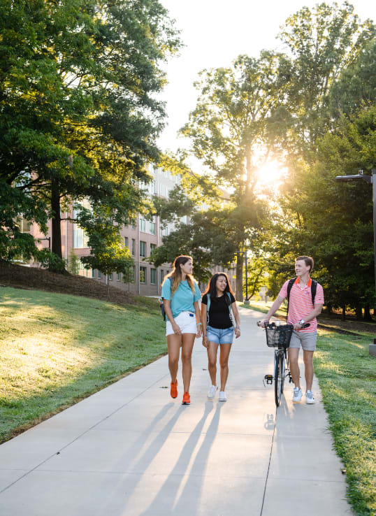 Three students walk beside Douthit Hills as the sun shines through the trees.