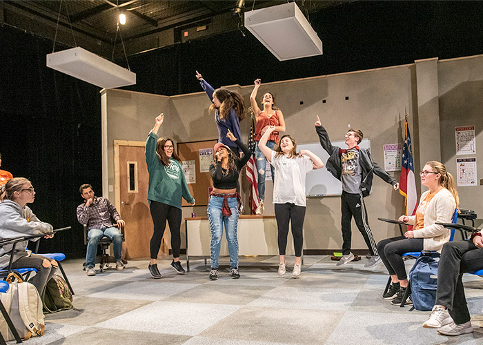 Six performing arts majors sing and jump in the air while on a set designed to emulate a classroom in the Brooks Center.