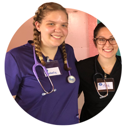 Student Isabelle Conrad, left, wearing purple medical scrubs and a name tag that says “Isabelle,” poses with a female student, right, wearing black medical scrubs.