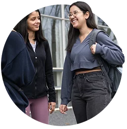 A female student wearing glasses, black jeans, and a blue top speaks with a female student wearing purple pants and a black jacket.