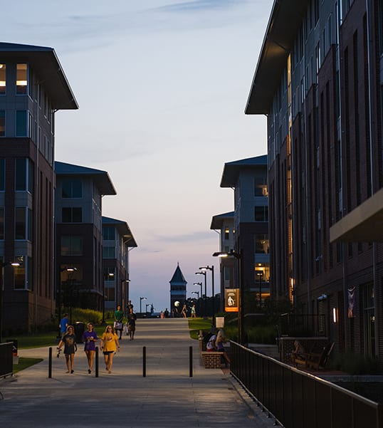 Clemson students walk between the buildings of Douthit Hills at dusk with Tillman Hall in the background.