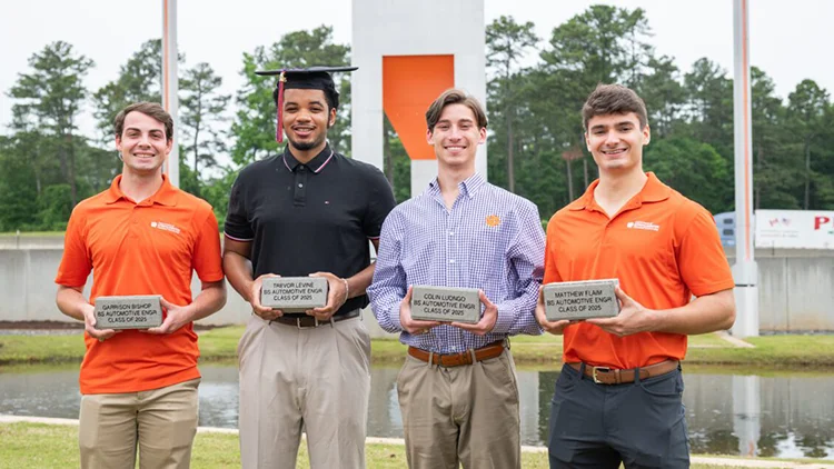 Four male students stand outdoors and hold commemorative bricks engraved
with their names, automotive engineering degree and 2025 graduation year. Two wear orange
Clemson University polos, one wears a black polo, and one wears a purple checkered shirt.