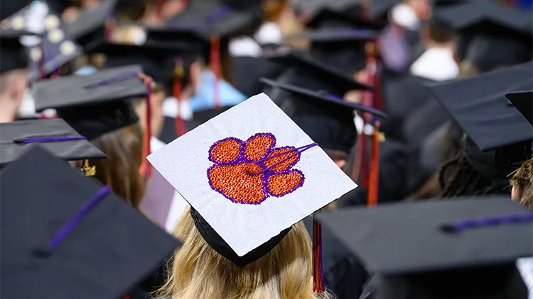 Graduation caps of students sitting in a graduation ceremony with a cap in center frame decorated with a Clemson Paw on a white background
