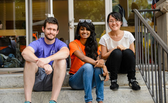 Two international female students sit with their male classmate and smile on the steps of Lee Hall during the spring.