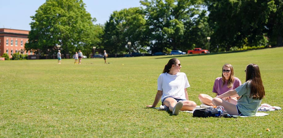 Three female undergraduate students smile and talk, sitting together in the sunshine on Bowman Field.