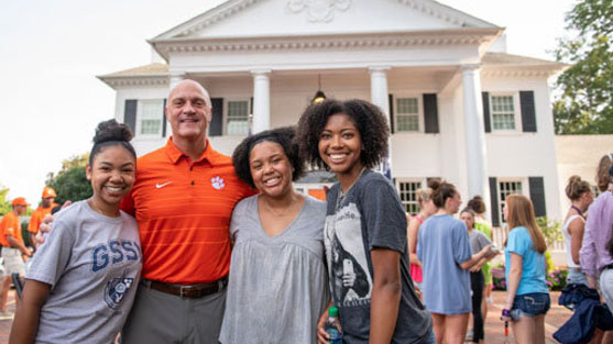 Three Black female students smile with President Clements on the front lawn of the President's Home during the freshman BBQ.