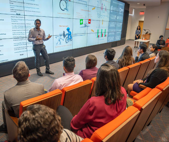 A male lecturer presents on a large screen to two rows of students in the Watt Innovation Center.