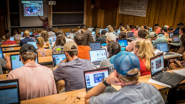 A classroom full of students sit behind their laptops in an auditorium while the instructor gives a presentation at the front