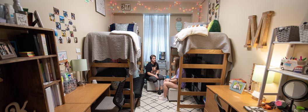 two female students sit on their beds in a decorated dorm room