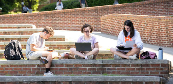 Two female and one male student study outside on a brick ledge on campus on a sunny day