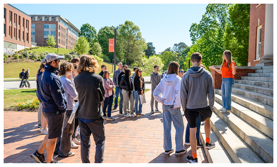 A female student stands on concrete steps and addresses a large crowd while giving a campus tour.