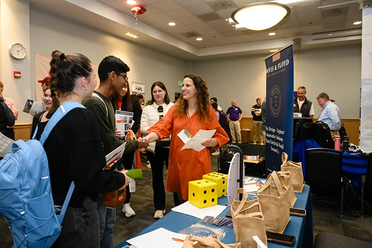 A female staff member wearing an orange dress shakes a male student's hand while standing in front of a table full of papers and small beige tote bags.