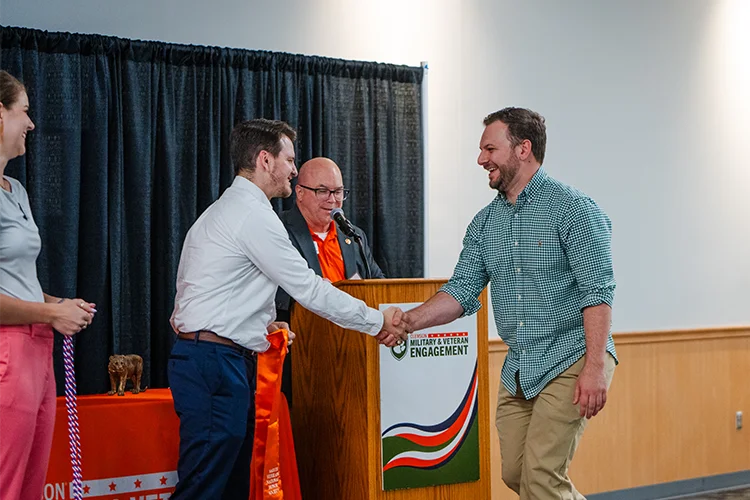 A man wearing khakis and a green gingham button-down shirt with rolled-up sleeves shakes the hand of a man wearing navy pants and a white button-down shirt while a man stands at a podium in the background and a woman holding a red, white and blue graduation cord stands to the side.