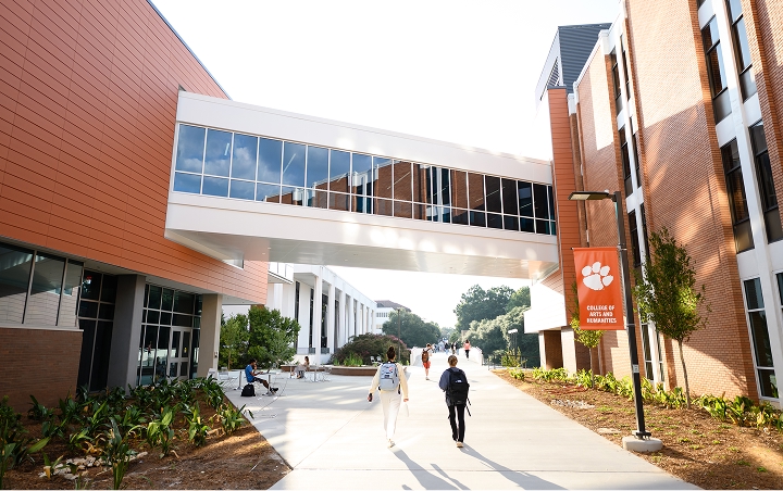 People walk on a sidewalk between two academic buildings connected by an elevated skywalk.