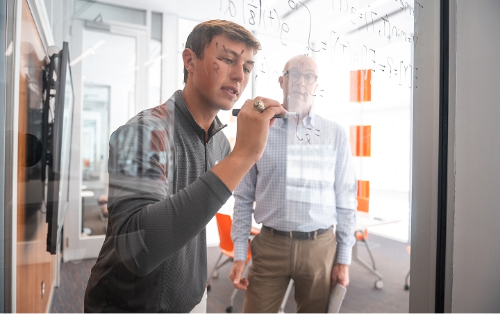 A male student writes mathematical equations on a clear wall as a male professor watches.