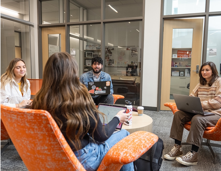 A male student and three female students sit in a circle in lounge chairs in a meeting room.