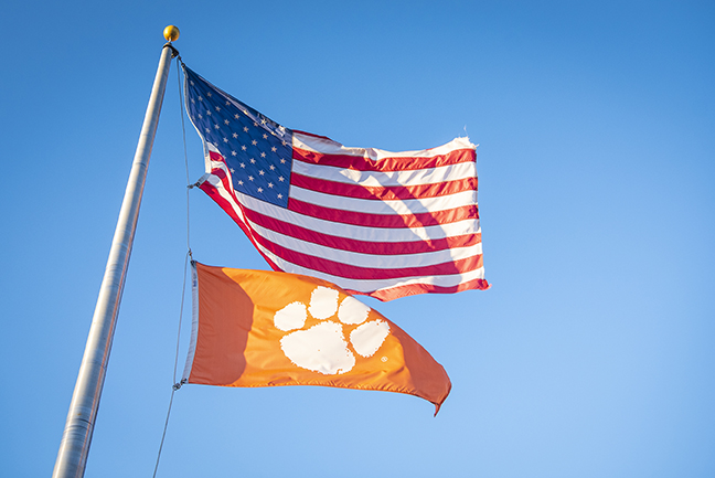 The American flag and a Clemson flag blowing in the wind on the same flag pole.