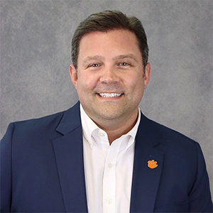 Headshot of a smiling man wearing a navy blazer and a white shirt, featuring a small orange floral emblem on the left lapel.