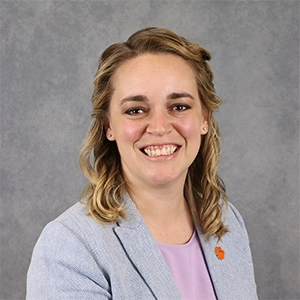 Portrait of a smiling woman with shoulder-length wavy hair, wearing a light gray blazer over a lavender top.