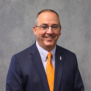 Professional portrait of a man in a navy suit and orange tie, smiling against a gray background.