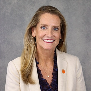 Professional headshot of a woman with long blonde hair, wearing a cream blazer and a dark blouse, smiling warmly against a textured gray background.