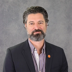 Professional headshot of a man with a beard wearing a black blazer and a checked shirt, against a gray background.