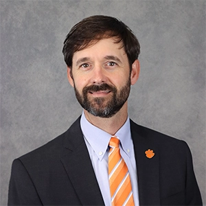 Professional headshot of a man in a suit with a Clemson University logo on his lapel, smiling against a gray background.