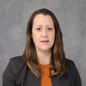 Professional headshot of a woman in a blazer with a subtle smile and shoulder-length hair against a textured gray background.