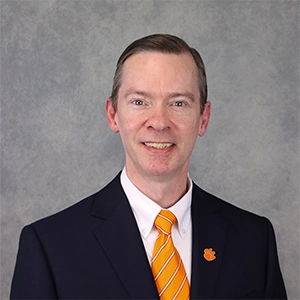 Formal headshot of a man in a suit with a Clemson University logo on his lapel, smiling against a neutral background.