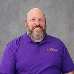 Headshot of a smiling man with a beard, wearing a purple polo shirt featuring the Clemson University logo.