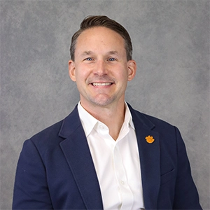 Professional headshot of a smiling man in a navy blazer and white shirt, featuring a distinctive orange lapel pin.