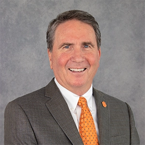 Professional headshot of a smiling man in a suit and tie, featuring a bright orange tie.
