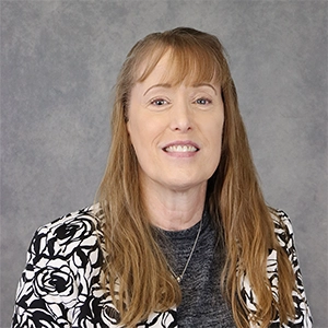 Professional headshot of a woman with long brown hair, wearing a black and white floral blazer.