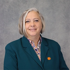 A professional portrait of a woman with short gray hair, wearing a teal blazer and a floral shirt, smiling confidently at the camera.