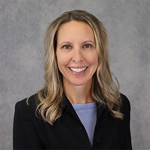 Professional headshot of a woman with long blonde hair, wearing a black blazer and smiling against a neutral gray background.