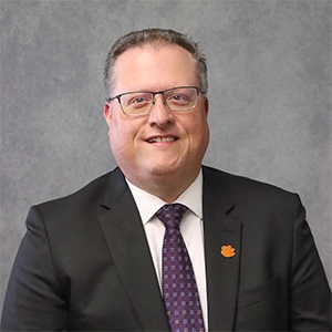 Professional portrait of a man in a suit with glasses, featuring a small orange Clemson University logo on his lapel.