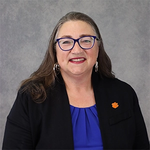 Professional portrait of a woman with long hair, wearing a blue top and dark blazer, smiling with glasses and an orange flower pin.