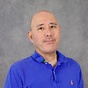 A headshot of a man with a bald head wearing a blue polo shirt, smiling softly against a gray background.