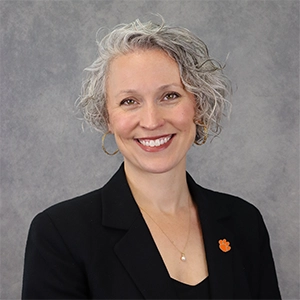 Headshot of a woman with short, curly gray hair, wearing a black blazer and a small orange paw print pin.